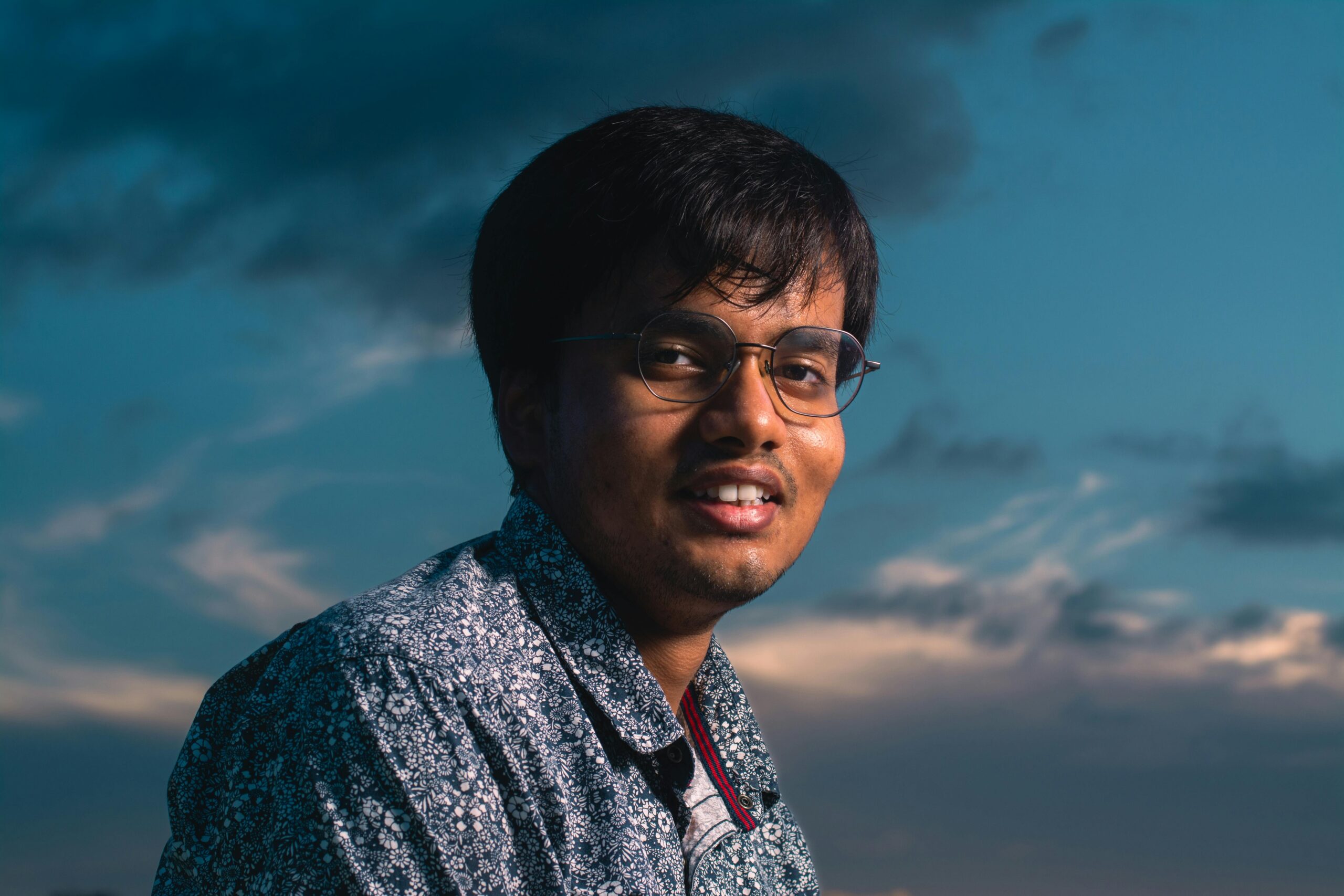 A smiling man in eyeglasses and a patterned shirt poses outdoors against a dynamic twilight sky.